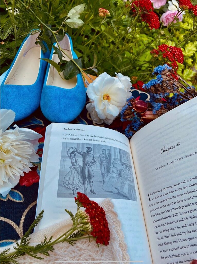 Bright blue shoes are positioned near an open book featuring an illustration, surrounded by fresh flowers. The scene conveys a cozy reading experience in a floral setting.